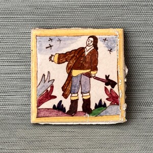 May include: A small, square, hand-painted tile with a historical illustration. The image depicts a person in a brown coat and boots holding an axe, with birds in the sky and colorful foliage. The tile has a yellow border.