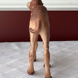 May include: A detailed figurine of a camel, viewed from the rear, showcasing its tan and brown coloration. The camel's legs are slender, and its tail is curled. The figurine is set against a neutral background.