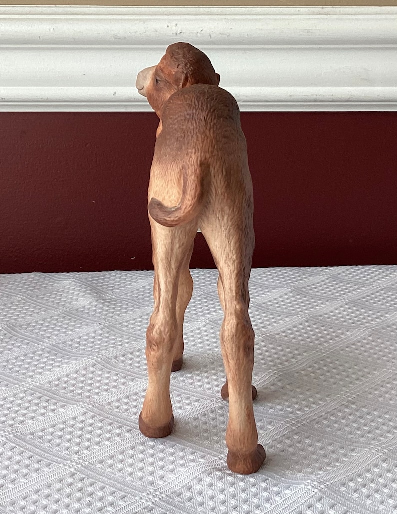 May include: A detailed figurine of a camel, viewed from the rear, showcasing its tan and brown coloration. The camel's legs are slender, and its tail is curled. The figurine is set against a neutral background.