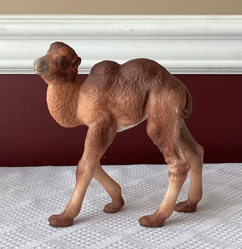 May include: A ceramic camel figurine in shades of brown and tan. The camel is posed with its head turned slightly to the left, and its legs are positioned as if walking. The figurine has a detailed texture and a smooth finish.