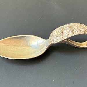 May include: A silver-toned spoon featuring an oval bowl and a curved, loop-shaped handle. The handle is decorated with a detailed, raised floral pattern that covers the entire surface of the loop, extending from the bowl to the end of the handle.