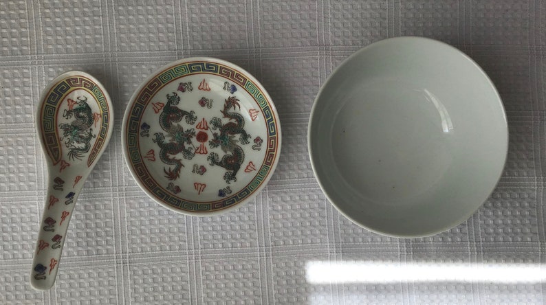 May include: A white ceramic bowl with a simple design, a white ceramic plate with a colorful dragon design, and a white ceramic spoon with a colorful dragon design.