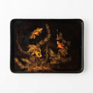 May include: A rectangular black serving tray featuring a hand-painted design of three orange goldfish swimming among golden aquatic plants and bubbles. The artwork is rendered in shades of orange, gold, and yellow against a dark background.
