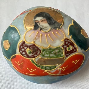 May include: A small, round, ceramic trinket box with a blue background and a painted image of a person in a colorful robe. The person is surrounded by gold, red, and green floral designs.