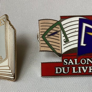 Op de afbeelding: Twee gouden spelden met boekontwerpen. De eerste speld heeft een gesloten boek met de tekst "APOSTROPHES PARIBAS". De tweede speld heeft een open boek met de tekst "SALON DU LIVRE".