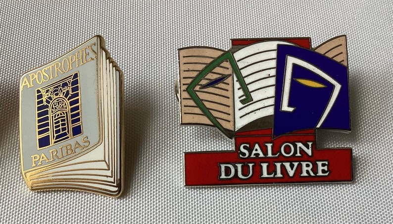 Op de afbeelding: Twee gouden spelden met boekontwerpen. De eerste speld heeft een gesloten boek met de tekst "APOSTROPHES PARIBAS". De tweede speld heeft een open boek met de tekst "SALON DU LIVRE".