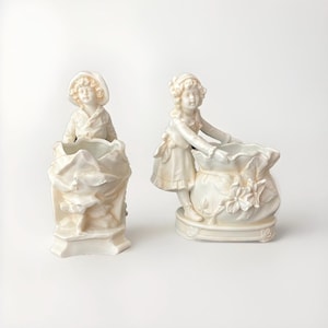 May include: Two white porcelain figurines of girls holding flower pots. The girl on the left is wearing a hat and a dress. The girl on the right is wearing a dress and holding a flower pot in the shape of a bag.