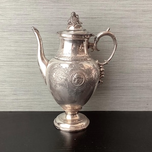 May include: A silver-colored antique coffee pot with ornate detailing. The pot features a curved spout, a handle, and a lid with a decorative finial. The body of the pot is engraved with floral and face designs, and it stands on a pedestal base.