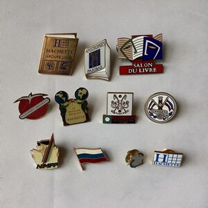 Op de afbeelding: Een verzameling van 11 kleurrijke emaille-pins met verschillende logo's en ontwerpen. De pins bevatten logo's voor Hachette, Salon du Livre, Passport Securite, Le Monde en Bleu en andere. De pins zijn allemaal in verschillende kleuren en stijlen en zouden een geweldige aanvulling zijn op elke verzameling.