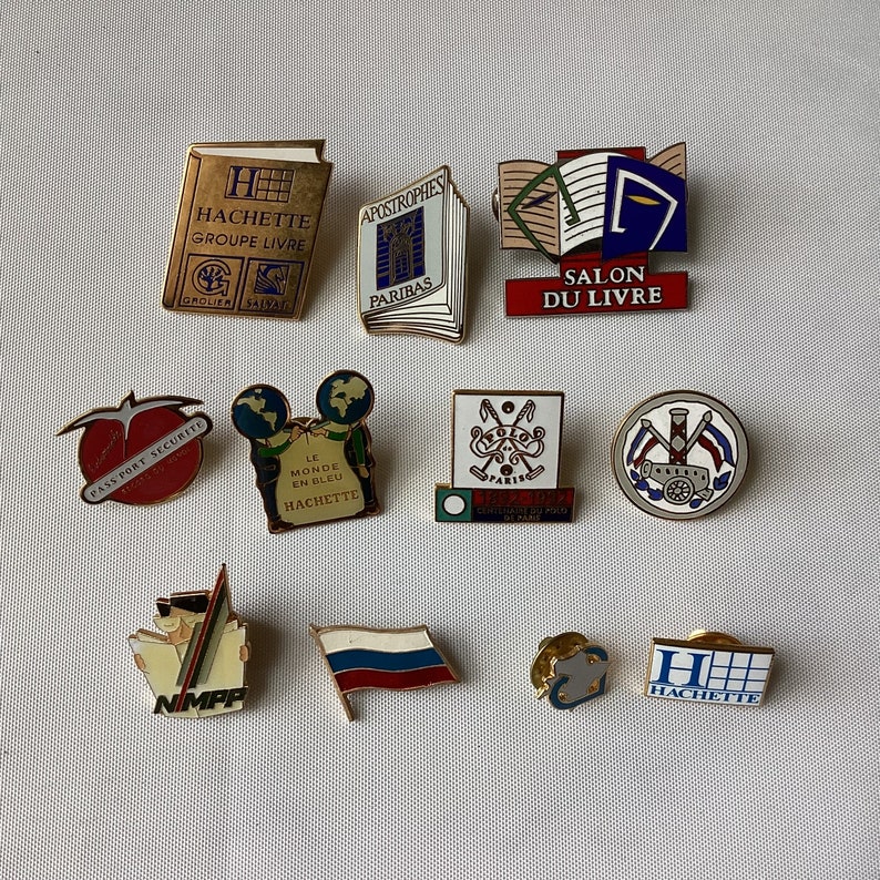 Op de afbeelding: Een verzameling van 11 kleurrijke emaille-pins met verschillende logo's en ontwerpen. De pins bevatten logo's voor Hachette, Salon du Livre, Passport Securite, Le Monde en Bleu en andere. De pins zijn allemaal in verschillende kleuren en stijlen en zouden een geweldige aanvulling zijn op elke verzameling.