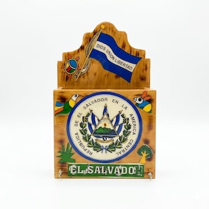 May include: A wooden wall-mounted mail holder featuring a painted blue flag with the text DIOS UNION LIBERTAD, the coat of arms of El Salvador with the text REPUBLICA DE EL SALVADOR EN LA AMERICA CENTRAL, and the text EL SALVADOR at the bottom.