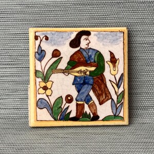 May include: A hand-painted ceramic tile featuring a person playing a lute in a garden setting. The tile has a yellow background with blue, green, and brown accents.