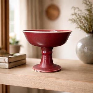 May include: A deep red ceramic pedestal bowl with a white rim. The bowl has a wide, shallow basin and a flared base. The bowl is likely used for serving fruit or displaying decorative items. The bowl is approximately 8 inches in diameter and 6 inches tall.