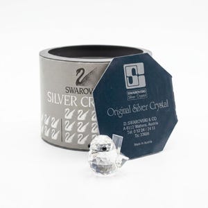 May include: A clear faceted crystal bird figurine with black eyes positioned in front of a cylindrical grey box and a dark blue hexagonal tag. The box features the Swarovski swan logo and text, while the tag displays the text Original Silver Crystal.