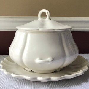 May include: White ceramic soup tureen with a lid and a matching plate. The tureen has a rounded, bulbous shape with a fluted design. The lid has a small handle on top.