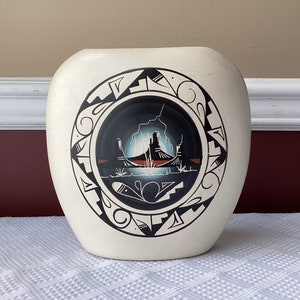 May include: White ceramic vase with a black and white geometric design featuring a stylized landscape scene. The vase has a rounded shape and a smooth finish.