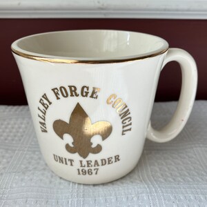 May include: White ceramic mug with a gold rim and gold lettering that reads "Valley Forge Council" and "Unit Leader 1967". A gold fleur-de-lis is in the center of the mug.