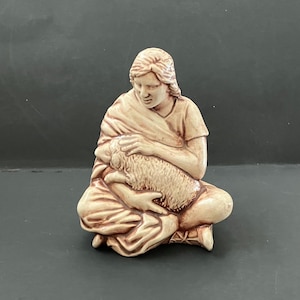 May include: A ceramic figurine depicting a person seated and cradling a lamb. The figure is dressed in a draped garment and has a serene expression. The lamb is light brown, and the figurine is a warm, earthy tone.