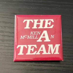 May include: A square, red button with white text. The text reads "THE KEN McMILLAN TEAM" in a bold, sans-serif font. The button is likely a promotional item or campaign button.