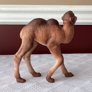 May include: A detailed figurine of a camel in a walking pose. The camel is primarily brown with lighter tan legs and a darker brown hump and head. The figurine is set against a neutral background.