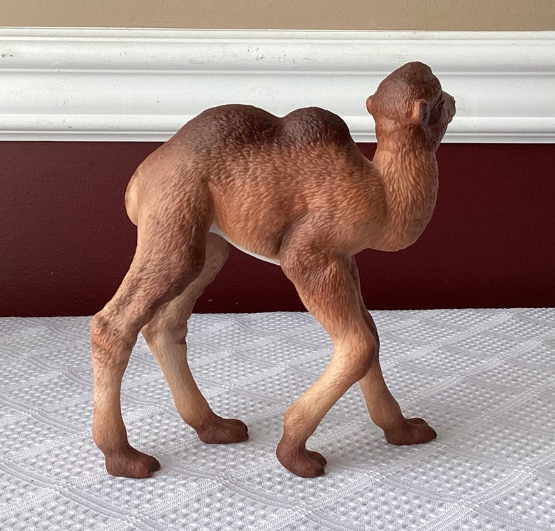 May include: A detailed figurine of a camel in a walking pose. The camel is primarily brown with lighter tan legs and a darker brown hump and head. The figurine is set against a neutral background.