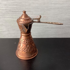 May include: A copper-colored Turkish coffee pot with a long handle and a lid. The pot has an ornate floral design on the lower portion and a spout for pouring. The handle is attached to the side of the pot.