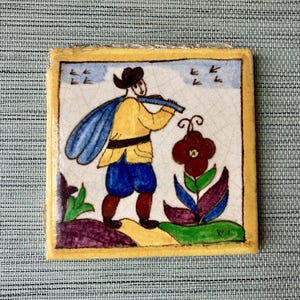 May include: Square ceramic tile with a hand-painted illustration. The image depicts a figure in a yellow jacket and blue shorts playing a flute, with a flower and birds in the background. The tile has a yellow border.