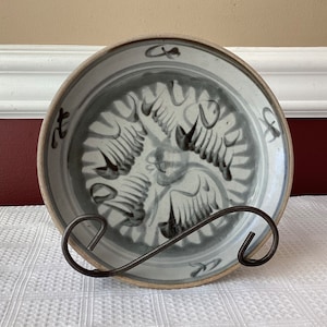 May include: A white ceramic plate with a blue and black design of stylized birds. The plate has a raised rim and is decorated with black calligraphy around the edge.