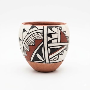 May include: A small, round, hand-painted ceramic pot with a terracotta base and rim. The white body features geometric designs in black and reddish-brown, including triangles, lines, and a mountain motif. The pot has a smooth, matte finish.