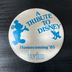 May include: A white button with blue text and graphics. The text reads "A Tribute to Disney" and "Homecoming '85". A blue silhouette of Mickey Mouse and an elephant are also featured. The text "wiu" is at the bottom.
