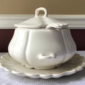 May include: White ceramic soup tureen with a lid and a matching plate. The tureen has a rounded, bulbous shape with a fluted design. The lid has a small handle on top.