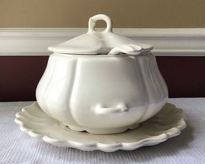 May include: White ceramic soup tureen with a lid and a matching plate. The tureen has a rounded, bulbous shape with a fluted design. The lid has a small handle on top.