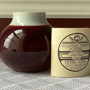 May include: A dark red and light blue ceramic vase with a glossy finish. The vase is sitting on a white tablecloth. A business card for Mountain Street Pottery is next to the vase. The card includes the address, phone number, and name of the business owner, Peter Jones.