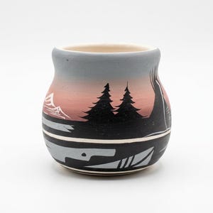 May include: A rounded ceramic pot featuring a matte finish with a gradient color scheme transitioning from light gray at the rim to a soft pink hue. The exterior is decorated with black silhouettes of pine trees, mountains, and geometric patterns.