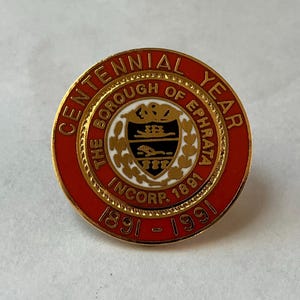 May include: A round, red and gold Centennial Year pin for The Borough of Ephrata, Inc. 1891-1991. The pin features a central emblem with a shield design and the text "The Borough of Ephrata" around the emblem.