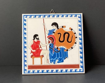May include: A ceramic tile with a white background and a blue, white, and orange border. The tile depicts two figures in a scene from Greek mythology. One figure is wearing a blue tunic and holding a shield with a snake design. The other figure is wearing a red tunic and holding a spear.