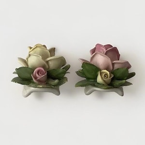 May include: Two porcelain rose bud figurines with green leaves. The roses are on small white bases. One rose is white and pink, the other is pink and yellow.