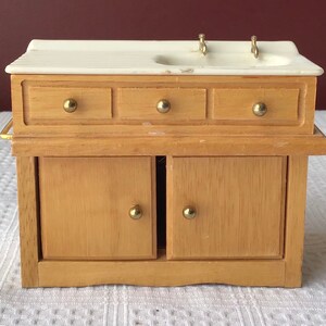 May include: A wooden dollhouse sink with a white porcelain countertop, two brass faucets, three drawers, and two cabinet doors with brass knobs.