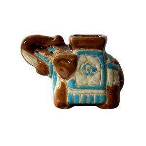May include: A ceramic elephant figurine in shades of brown, white, and turquoise. The elephant has a decorative saddle with floral details and a square opening on its back. The figurine is a decorative item.