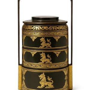 May include: A black and gold tiered food carrier with a handle. The carrier features ornate gold detailing and a mythical creature design. The handle and frame are black, and the carrier is approximately 10 inches tall.