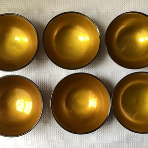 May include: Six gold-coloured bowls with black rims. The bowls are arranged in two rows of three. The bowls are on a white surface.