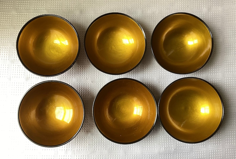 May include: Six gold-coloured bowls with black rims. The bowls are arranged in two rows of three. The bowls are on a white surface.