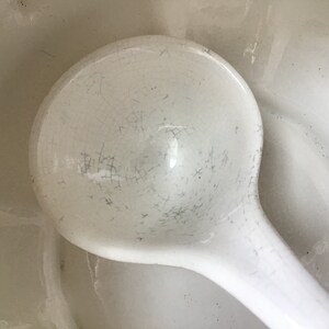 May include: A white ceramic ladle with a crazed glaze. The ladle is in a white ceramic bowl.