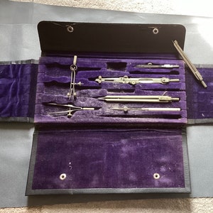 May include: A black leather case with a purple velvet interior holds a set of drafting tools, including compasses and rulers.