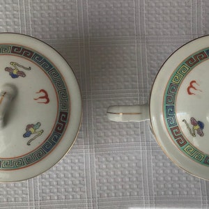 May include: Two white ceramic teacups with lids. The lids have a gold trim and a small knob on top. The cups are decorated with a blue and red geometric pattern and a stylized bird design.