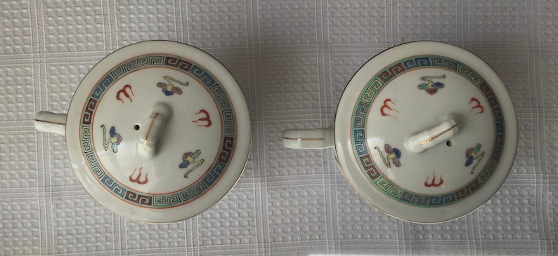 May include: Two white ceramic teacups with lids. The lids have a gold trim and a small knob on top. The cups are decorated with a blue and red geometric pattern and a stylized bird design.