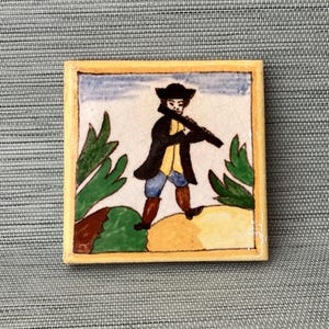 May include: A hand-painted ceramic tile featuring a man in a black coat and hat playing a flute. The tile has a yellow border and a white background with green foliage and brown hills.