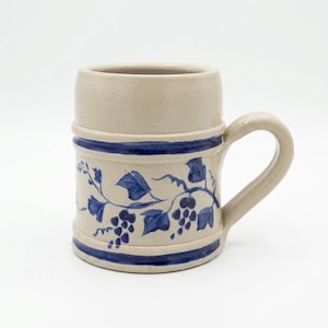 May include: A beige ceramic beer mug featuring a thick blue horizontal band at the top and bottom. The center of the mug is decorated with a hand-painted blue vine pattern that includes detailed grape clusters and leaves wrapping around the body.