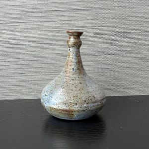 May include: A handcrafted ceramic vase with a bulbous body and a narrow neck. The vase features a speckled, textured surface with a blend of colors, including brown, blue, and beige. The vase is suitable for displaying flowers or as a decorative accent.