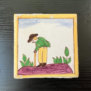 May include: A hand-painted ceramic tile with a yellow border depicting a cartoon figure in a green shirt and brown hat walking with a stick. The figure is standing on a purple hill with green plants.